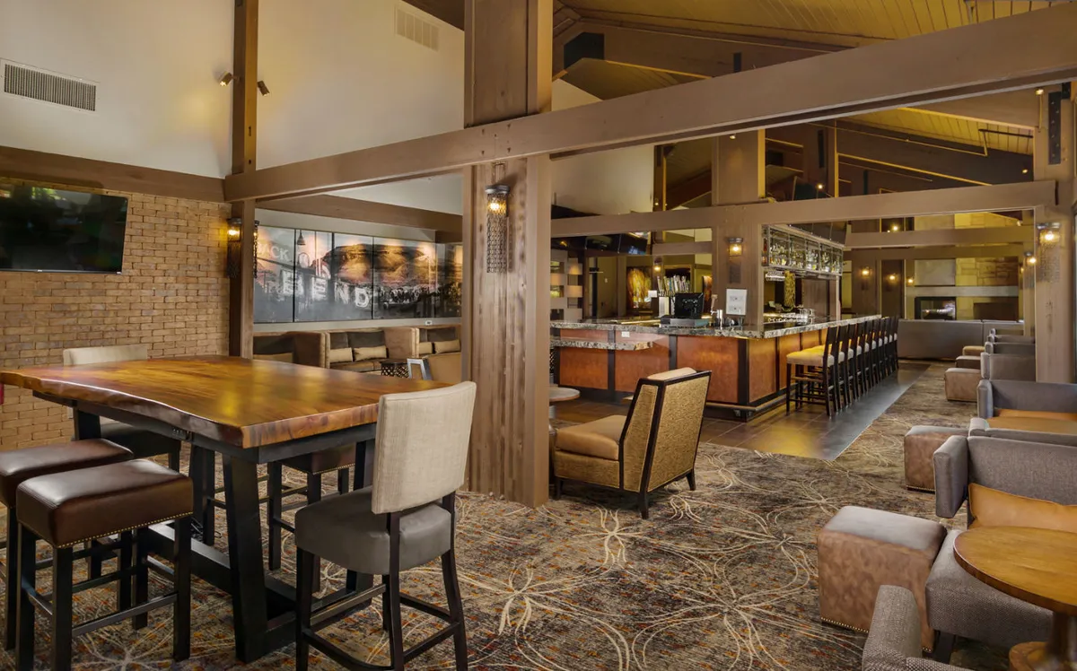 This interior image showcases a large wooden table surrounded by comfortable seating, a prominent bar area, and inviting decorative lighting. The arrangement suggests a lively yet cozy dining environment, perfect for group gatherings, shared meals, and socializing over drinks. LCD TVs are subtly visible, catering to sports fans as described in the section.