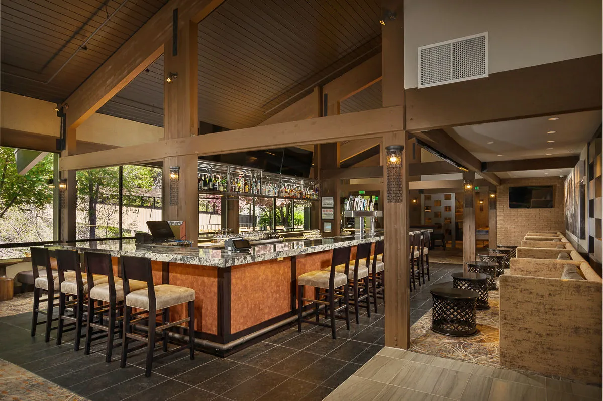 A vibrant lounge interior featuring a well-lit bar counter lined with stylish stools. Bottles of various spirits are attractively displayed behind the bar, and comfortable seating areas invite guests to relax. People are socializing over drinks, with several watching a sporting event on one of the numerous wall-mounted LCD TVs. The atmosphere is lively, friendly, and perfect for enjoying cocktails or a casual meal.