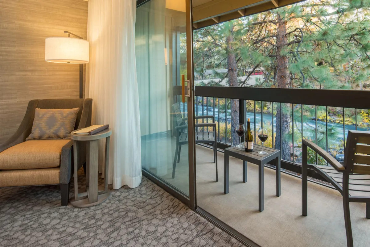 This interior shot shows a comfortable armchair beside a small table with wine glasses, facing a large glass door that leads out to a private balcony. Beyond the door is a glimpse of the natural scenery, hinting at river views and the opportunity to enjoy fresh air from your own outdoor space.