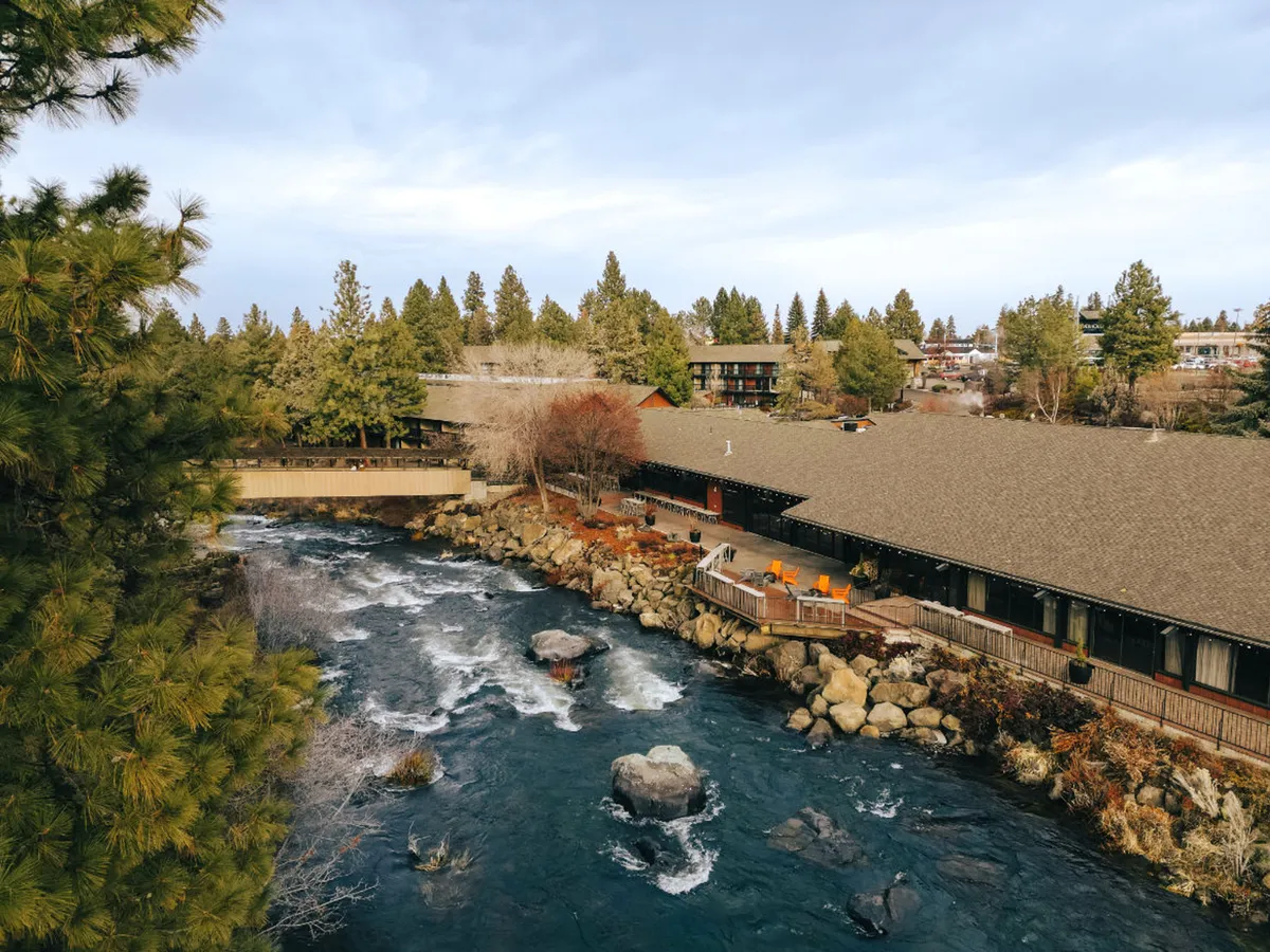 A serene shot of the Deschutes River with a spacious deck area in the foreground, surrounded by tall trees and the building's architecture partially visible. The scene captures people relaxing by the river, enjoying meals on the deck, and hints of nearby nature trails. The lighting suggests either early morning or sunset, adding to the tranquility.