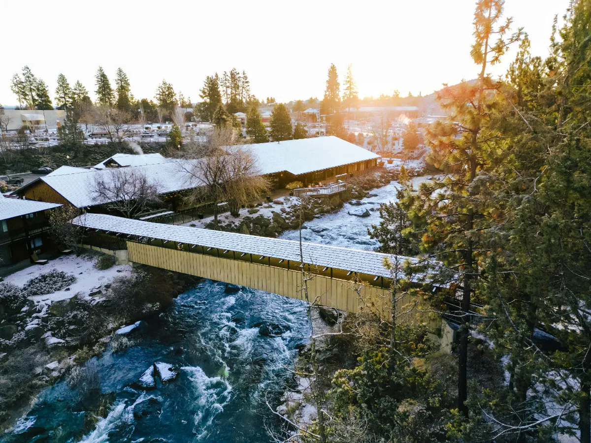 A tranquil exterior scene featuring a charming covered bridge spanning the flowing Deschutes River, surrounded by lush trees. There are people seen walking across the bridge, taking photos, and enjoying the natural surroundings. Buildings in the background reinforce the proximity to accommodations.