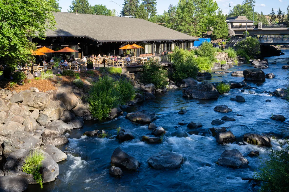 The image showcases an inviting restaurant patio with vibrant orange umbrellas perched alongside the gently flowing Deschutes River. The patio is set against a backdrop of lush greenery and a rocky riverbank, inviting guests to dine outdoors, unwind with friends, and soak in the tranquility of nature at sunset.