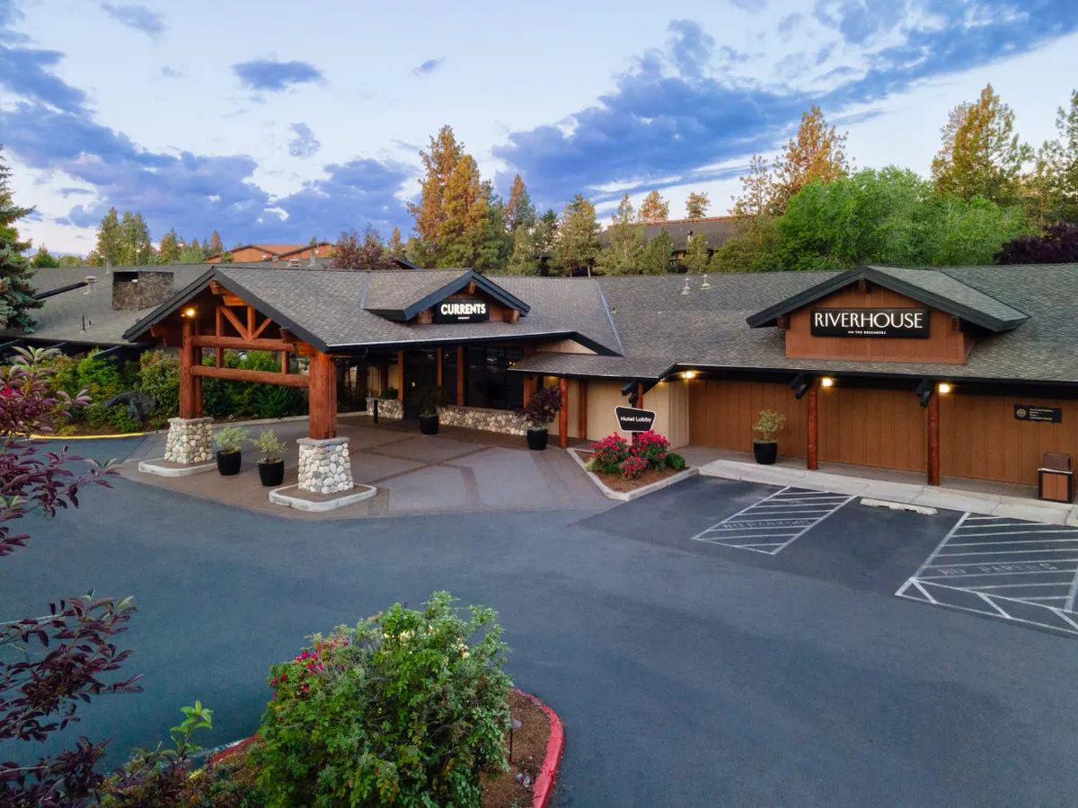 An inviting entrance scene with prominent wooden beams and well-maintained landscaping. Signs for 'Currents' and 'RiverHouse' are visible, with the entrance bathed in warm evening light. The surrounding greenery suggests proximity to nature trails, and there’s a sense of arrival and anticipation for further exploration.