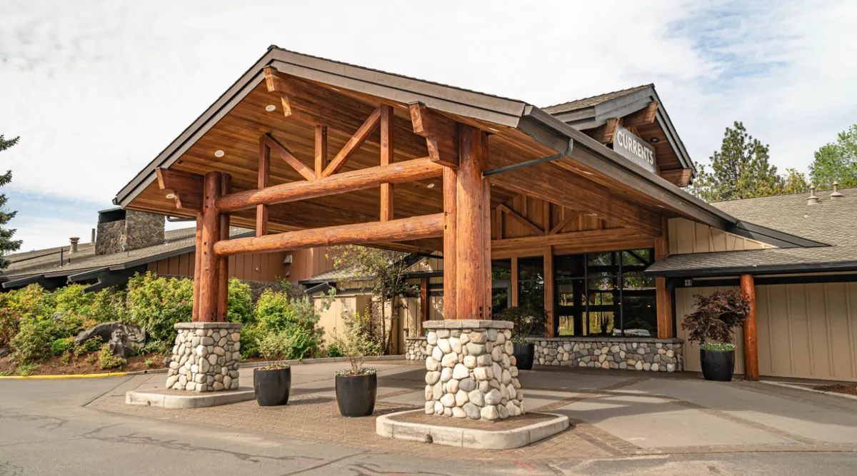 This image depicts the resort’s welcoming exterior, featuring large wooden beams, stone pillars, expansive glass windows, and beautifully landscaped greenery. Guests gather for events and enjoy the inviting atmosphere under the canopy.