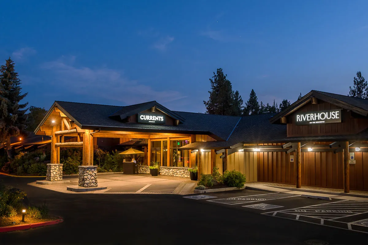 The exterior view highlights the entrance to the CURRENTS Restaurant and Lounge, with wooden structures, illuminated signage, and a landscaped area. The evening setting evokes warmth, social gatherings, and the promise of Northwest dining experiences.